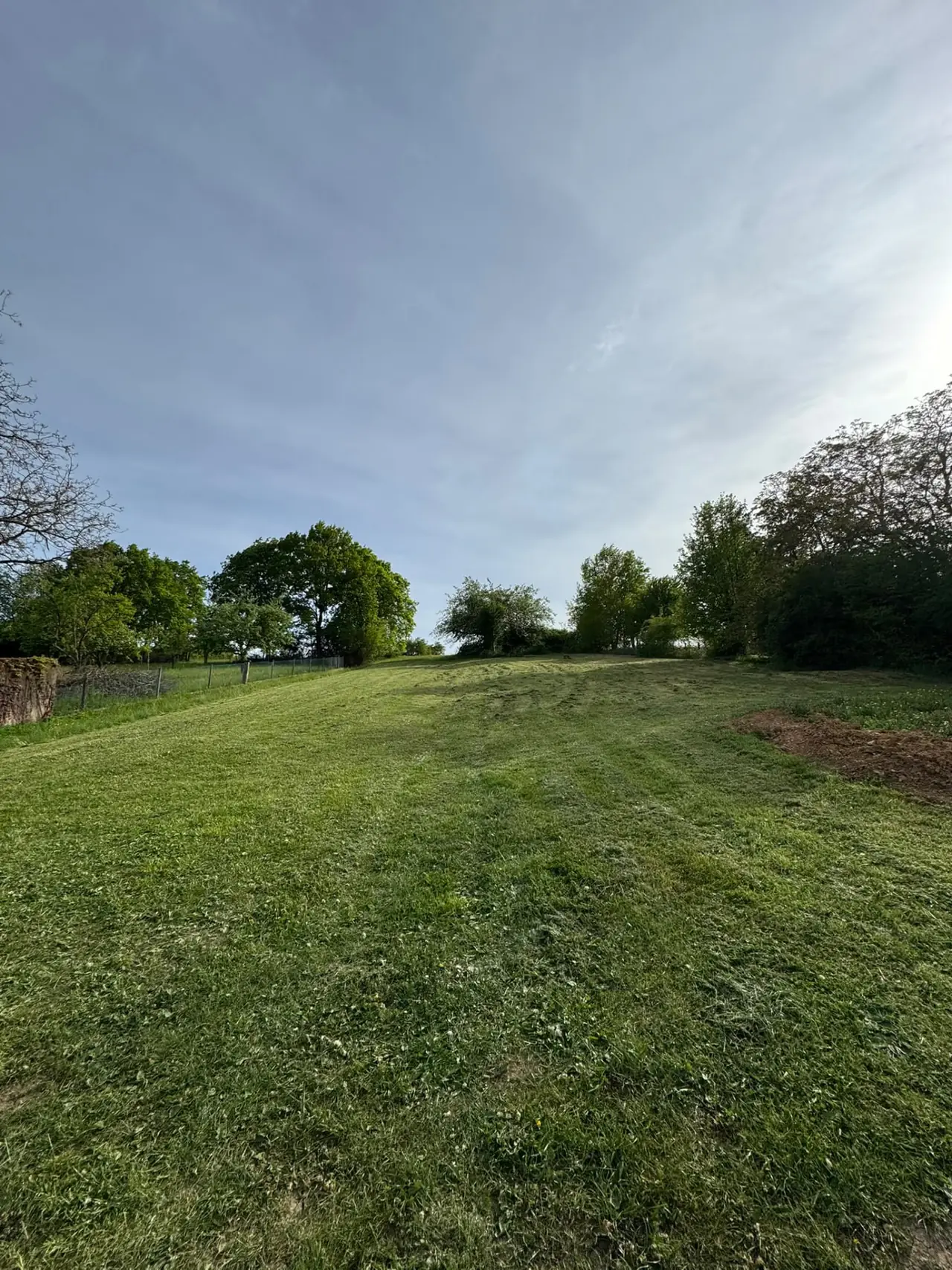 Terrain en pelouse en pente douce, bordé d’arbres et d’un grillage, avec un tas de terre sur le côté