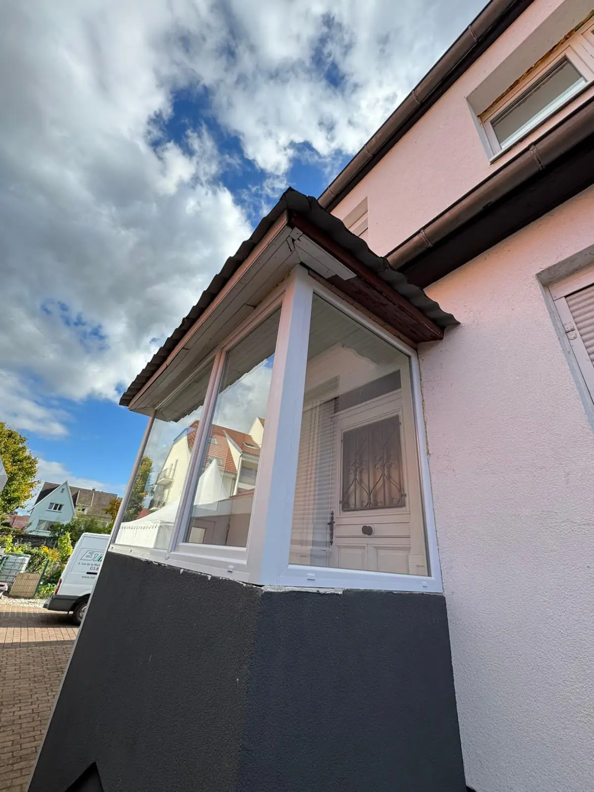 Avancée vitrée à menuiseries blanches sur soubassement anthracite, devant une porte d’entrée ancienne à grille ouvragée