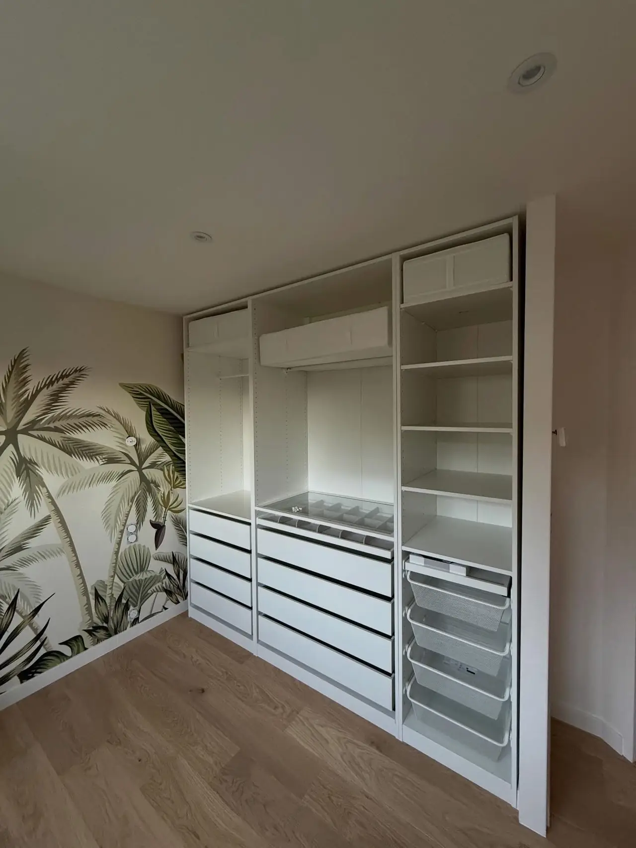 Rangement blanc posé contre le mur : penderie, étagères et tiroirs ; papier peint végétal sur le mur voisin, parquet au sol